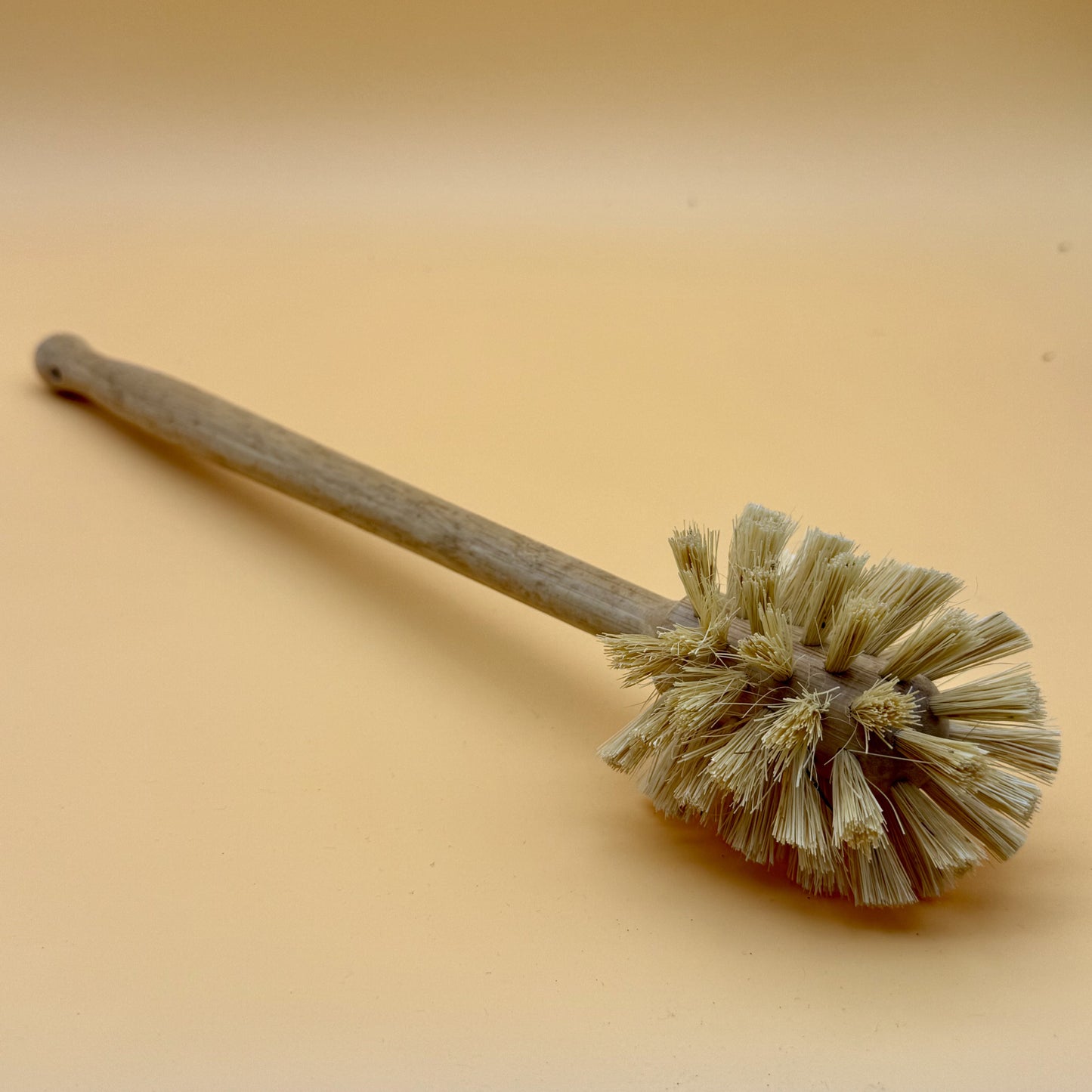 A rustic wooden cleaning brush with natural beige bristles, shown diagonally on a smooth, warm beige background.